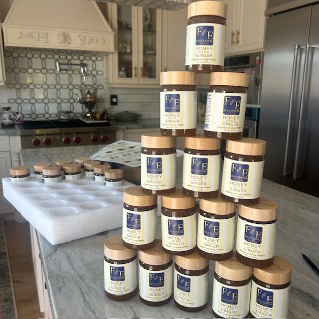 A pyramid of Functional Elixirs Honey with Fresh Ginger jars with wooden lids stacked on a marble kitchen counter